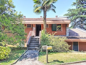 A two-story building with a stone staircase leading to the entrance, surrounded by lush greenery and a palm tree. - Hermitage by Interhome (Porto Valtravaglia)