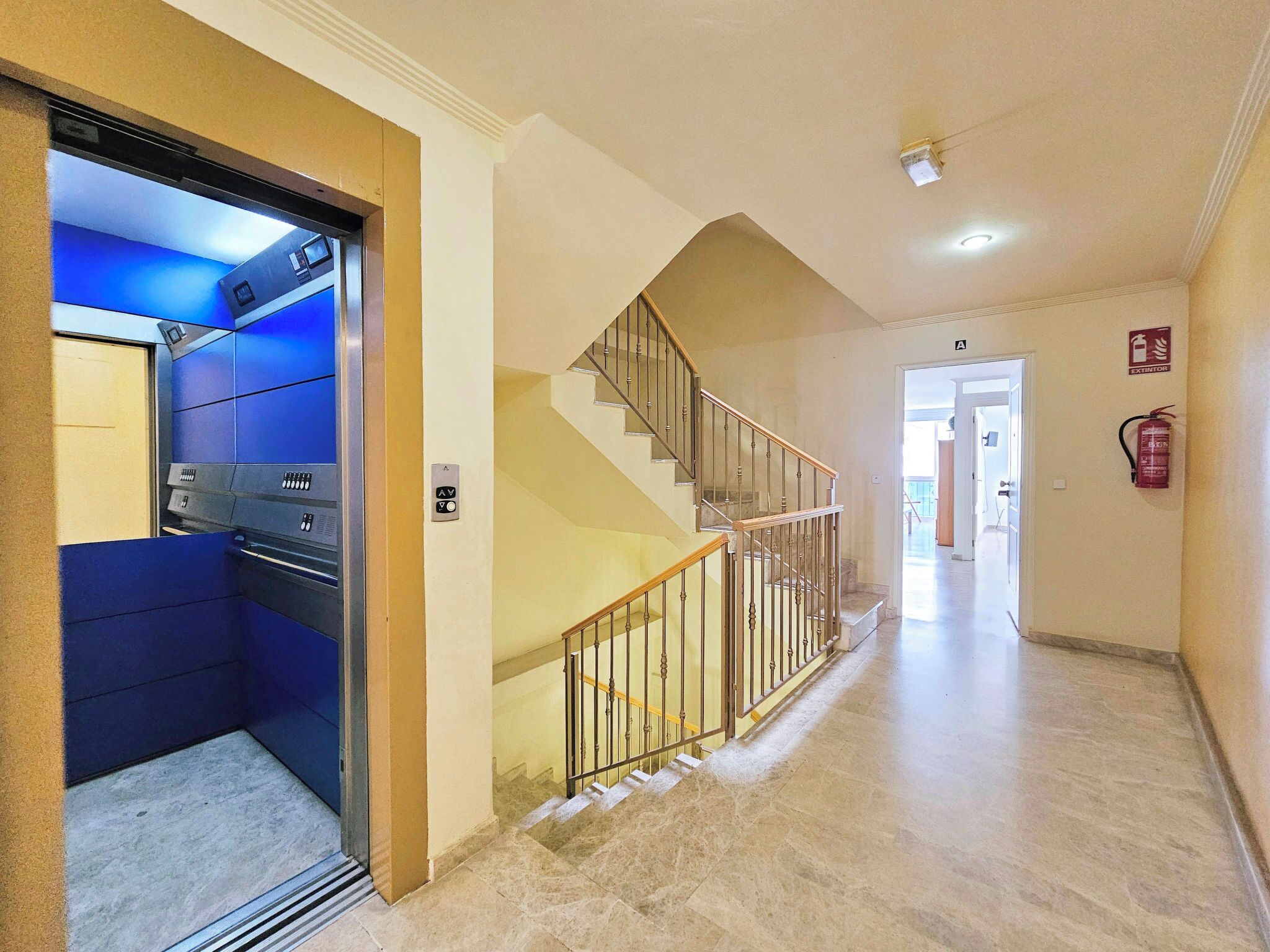 A hotel hallway with a blue elevator, a staircase, and a fire extinguisher.