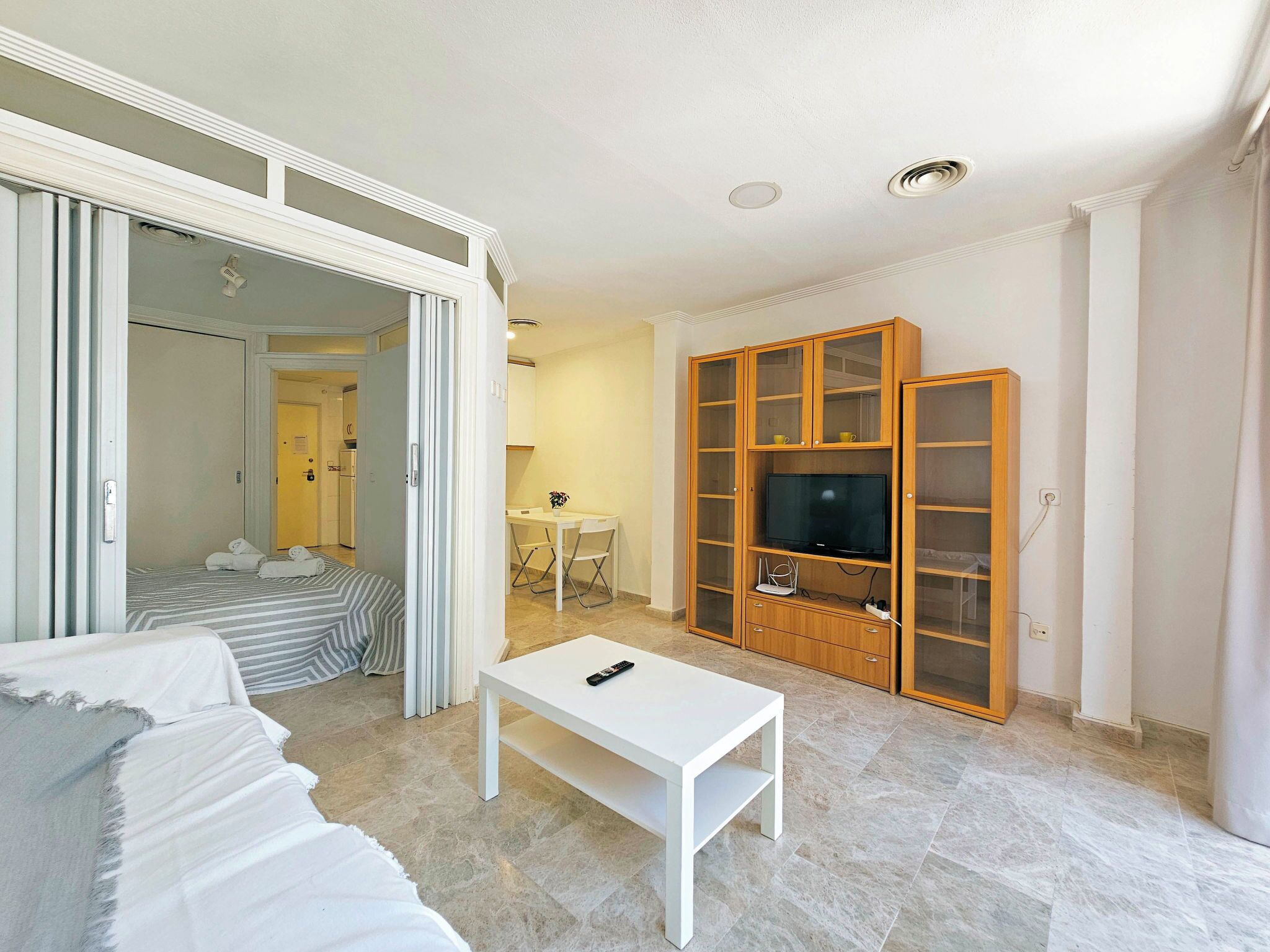A hotel room with a bed, a white sofa, a TV stand with a flat-screen TV, and a sliding door leading to a bathroom.