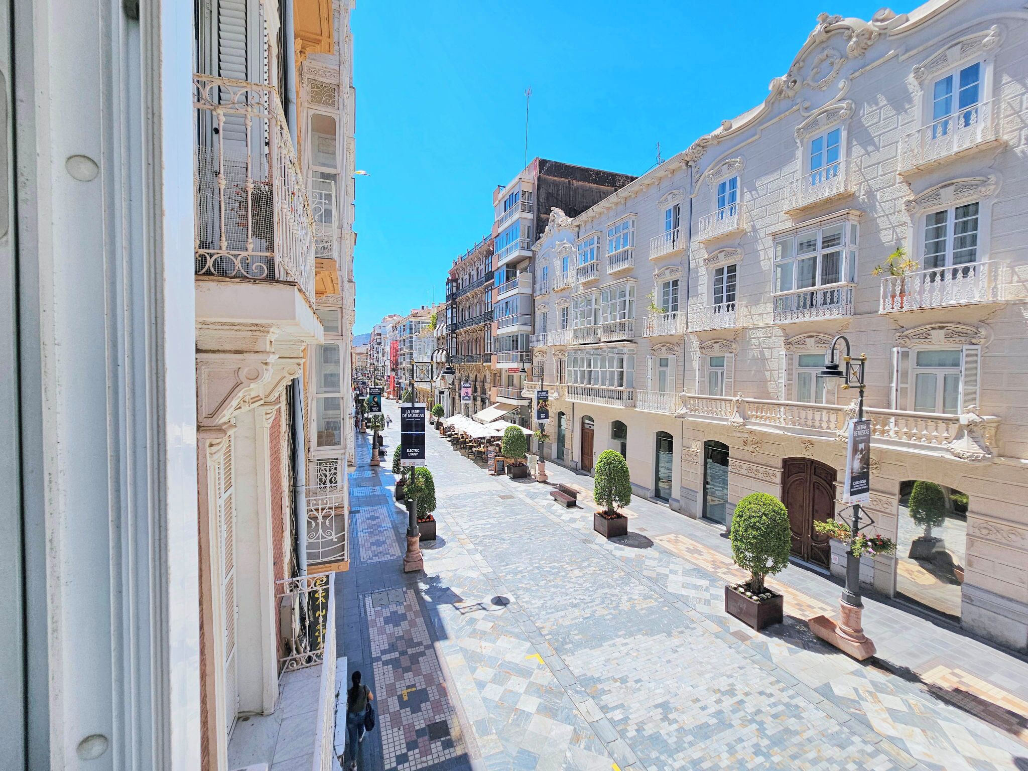 Une rue bordée d’immeubles, avec un trottoir pavé et des plantes en pot.