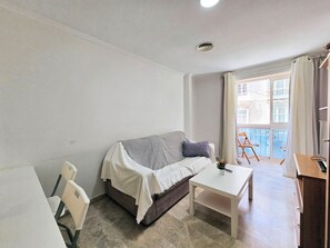 Ett litet, väl upplyst rum med en soffa, ett soffbord och en stol. Det finns ett fönster med gardiner och utsikt över byggnader utanför.