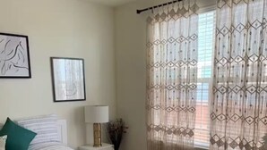 Quarto com cama, mesa de cabeceira, abajur e janela com cortinas estampadas.