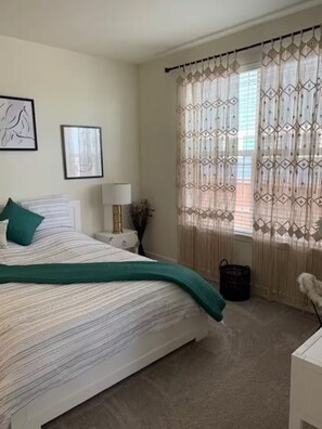 Ein Schlafzimmer mit Bett, Nachttisch, Lampe und einem Fenster mit gemusterten Vorhängen.