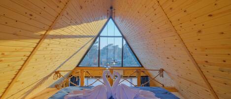 A triangular wooden room with a bed and a large window.
