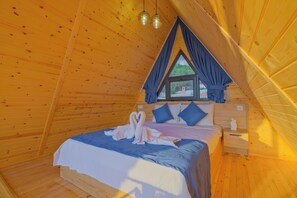 Una habitación acogedora de madera con una cama, almohadas, una mesita de noche y un ventanal con cortinas.