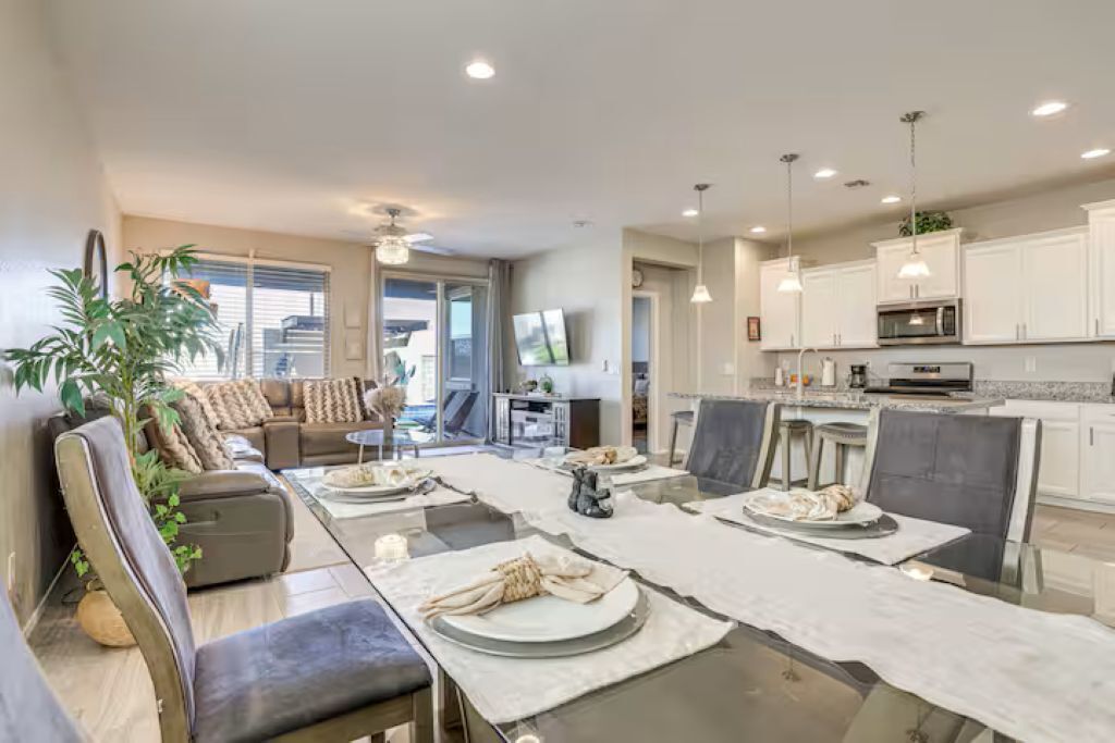 A modern living room with a dining area, a kitchen with white cabinets, and a large window providing natural light.