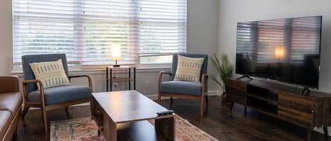 A living room with a sofa, two armchairs, a coffee table, a TV, and a window with blinds.