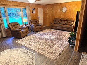 Un salon avec un grand tapis, deux fauteuils inclinables, un canapé et un mur en bois orné d’une horloge.