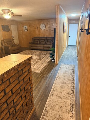 Un salon avec un mur d’accent en briques, un ventilateur de plafond, un canapé en cuir marron et un tapis à motifs.