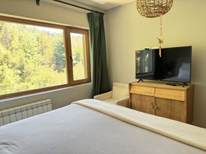 A bedroom with a bed, a wooden dresser, a television, and a window with a view of trees. - House of Worship (San Carlos de Bariloche)