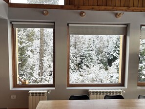 A room with a wooden table and two windows with wooden frames, each with a view of snow-covered trees. - House of Worship (San Carlos de Bariloche)