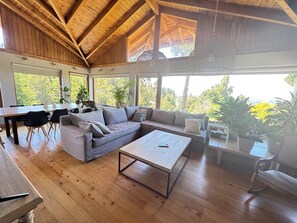 Ein großzügiger Wohnbereich mit Holzbalkendecke, großen Fenstern, einer Sofagarnitur, einem Couchtisch und einem Esstisch mit Stühlen.