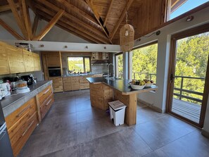 A modern kitchen with wooden cabinets, a large island, and a view of trees through the windows. - House of Worship (San Carlos de Bariloche)