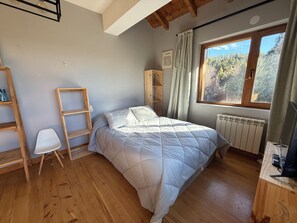 Ein Schlafzimmer mit einem Bett, einem Heizkörper, einem Fenster mit Blick auf Bäume, einem Schreibtisch mit Computer und einem Stuhl.