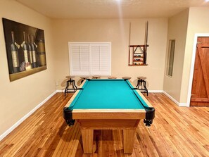 Una mesa de billar con superficie de juego color aguamarina, rodeada de taburetes de madera, en una habitación con piso de madera y un cuadro en la pared.