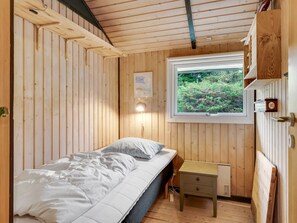 A compact bedroom with a single bed, a small bedside table, a window with a view of greenery, and wooden paneling on the walls.