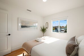 Ein Schlafzimmer mit einem Bett, einem Fenster mit Blick auf ein Haus, einem gerahmten Bild an der Wand und einer Pflanze.