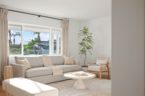 Ein modernes Wohnzimmer mit einer Couch, einem Sessel, einem Couchtisch und einem großen Fenster mit Vorhängen.