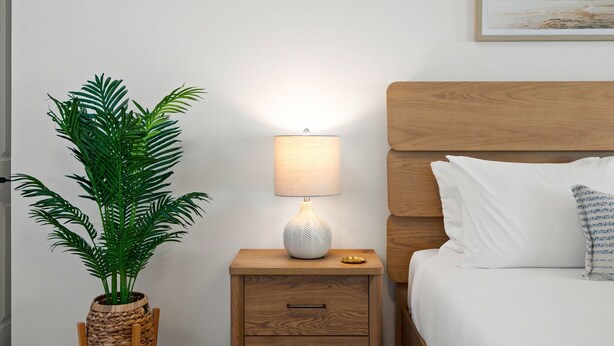 Ein ordentlich eingerichtetes Schlafzimmer mit einem hölzernen Kopfende, einem Nachttisch mit Lampe, einem Topf mit Pflanze und einem Bett mit Kissen.