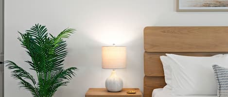 A neatly arranged bedroom with a wooden headboard, a bedside table with a lamp, a potted plant, and a bed with pillows.