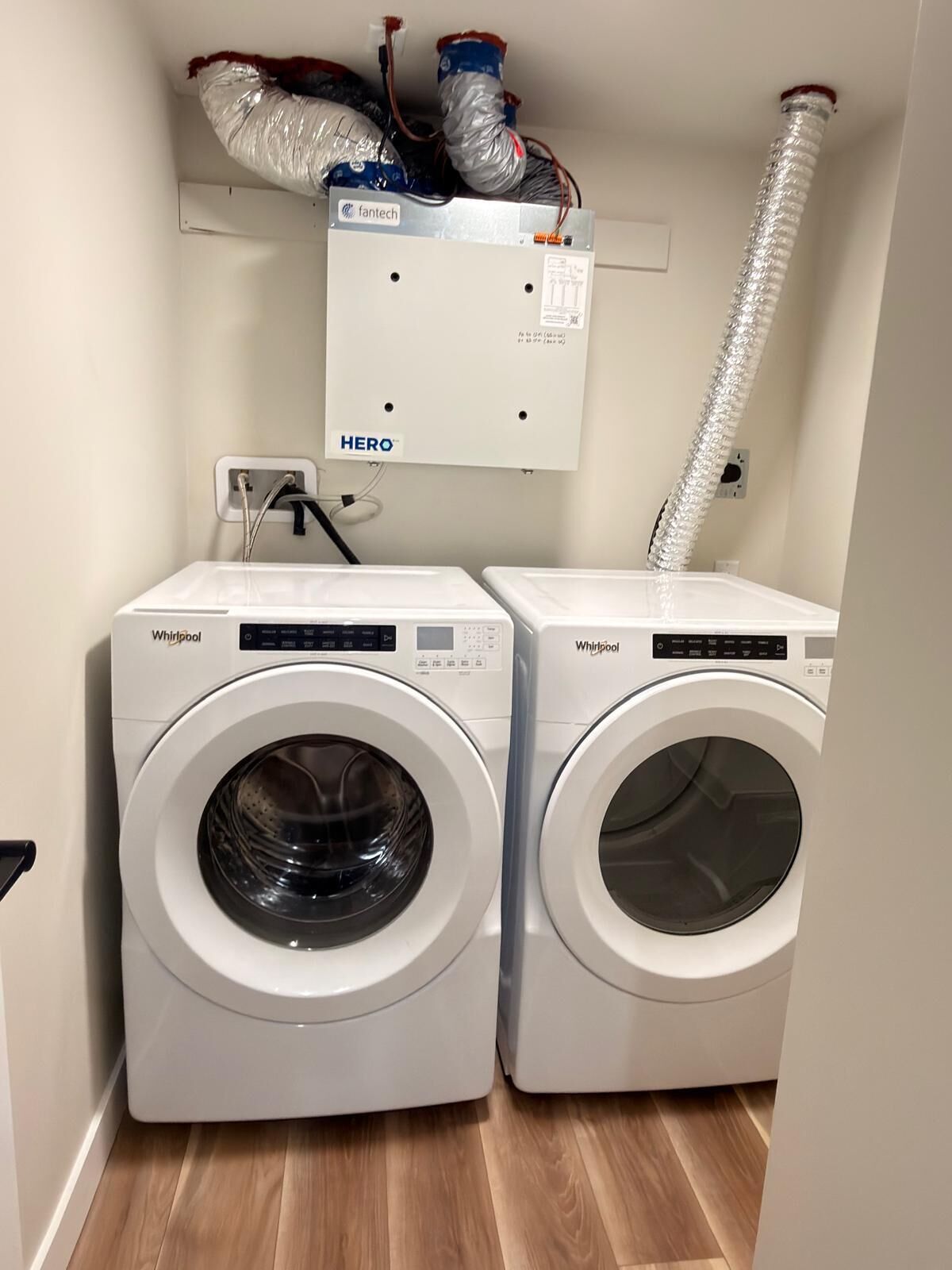Zwei Whirlpool Waschmaschinen mit einer weißen, an der Wand befestigten Bedienkonsole darüber, einem flexiblen Ablaufschlauch und einer Steckdose.