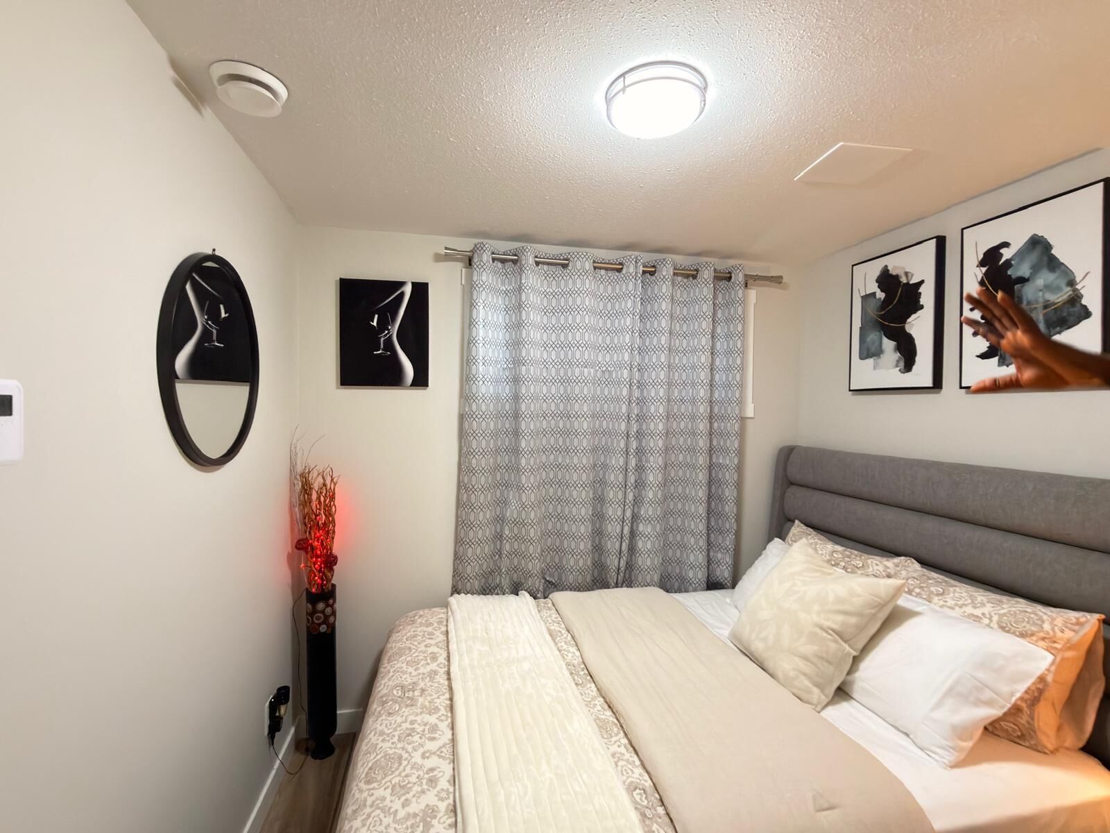 Une chambre à coucher comprenant un lit, une table de chevet, une horloge murale, une fenêtre avec des rideaux à motifs et des œuvres d’art encadrées accrochées au mur.