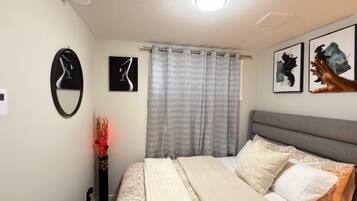 A bedroom with a bed, a bedside table, a wall-mounted clock, a window with patterned curtains, and framed artwork on the wall.