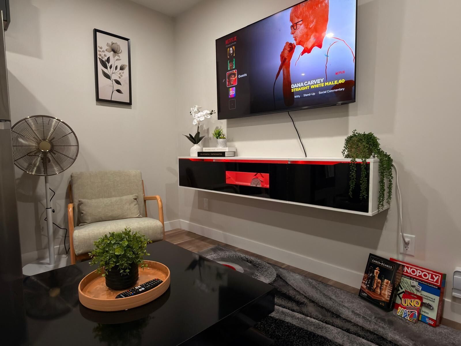 A modern living room with a flat-screen TV mounted on the wall, a sofa, a coffee table with a plant, and a fan.