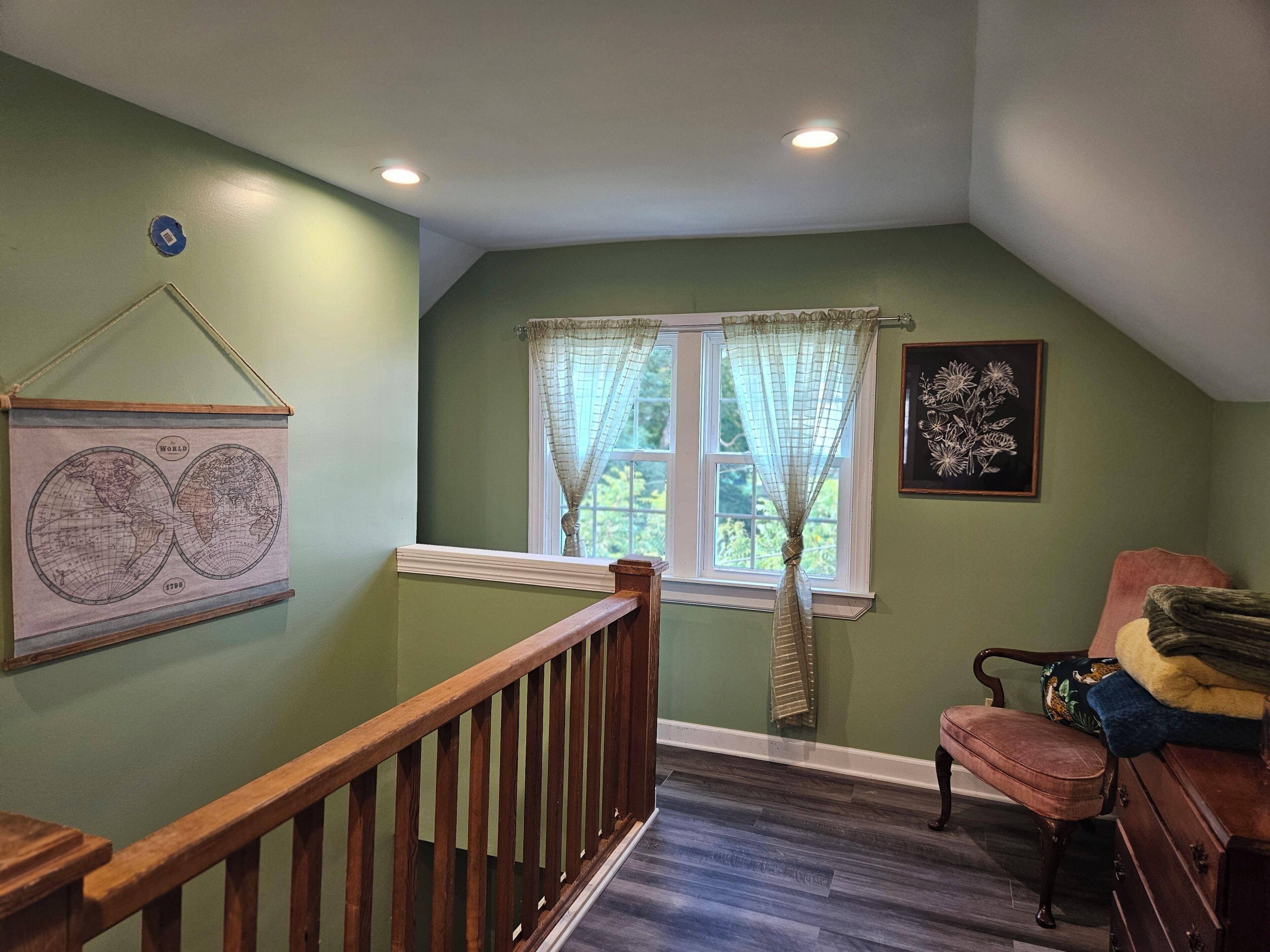 A room with a wooden staircase, a window with sheer curtains, a world map on the wall, and a floral artwork.