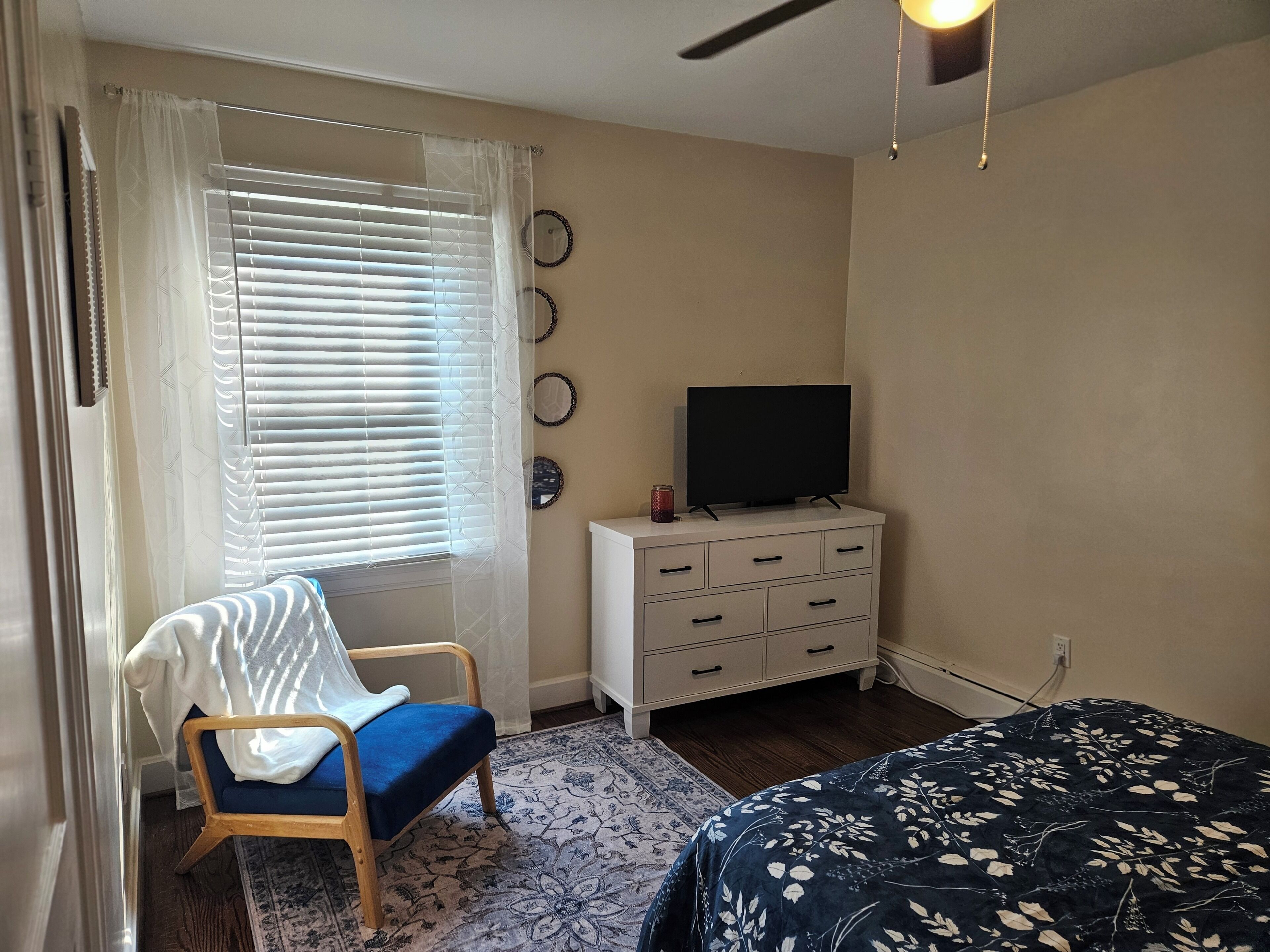 A bedroom with a bed, a chair, a TV on a dresser, and a window with blinds.