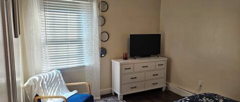 A bedroom with a bed, a chair, a TV on a dresser, and a window with blinds.