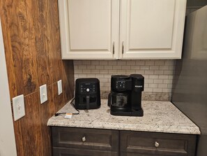 Une cuisine équipée d’une machine à café et d’une machine Keurig posées sur un plan de travail en granit, avec des armoires en bois et un dosseret de crédence en carrelage blanc.