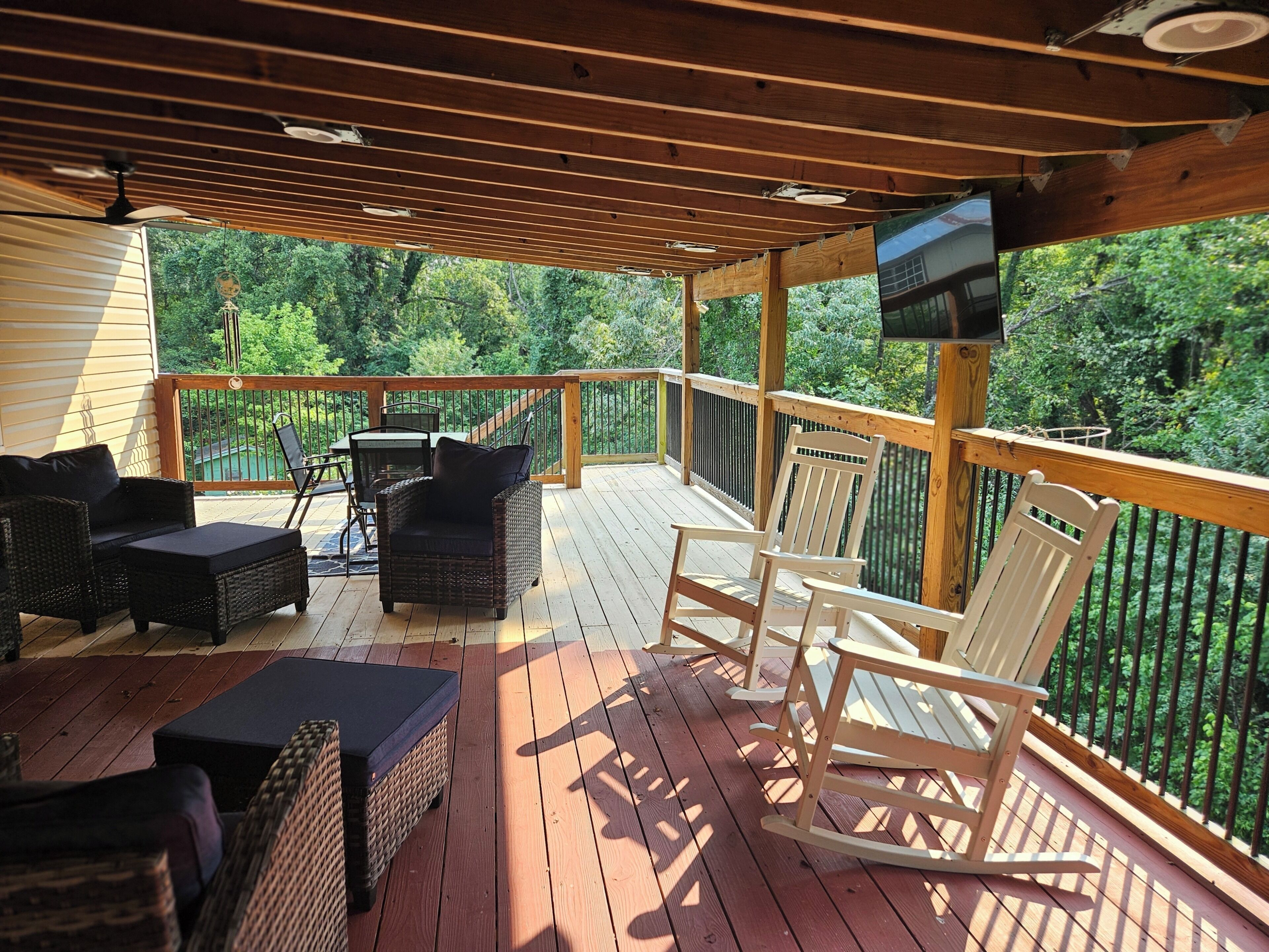 A covered deck with outdoor furniture, a television mounted on the wall, and a view of the surrounding greenery.