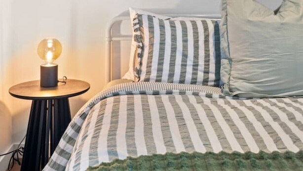 Uma cama com roupa de cama listrada, um abajur e uma luminária na parede.