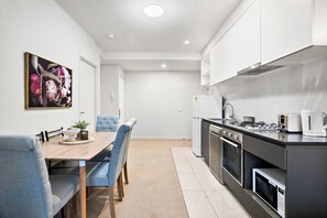 A modern kitchen with stainless steel appliances, a dining area with a wooden table and blue chairs, and a painting on the wall. - Collins St CBD escape| Pool, Gym, Sauna & Parking (Melbourne)