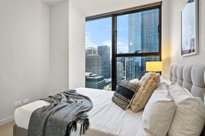 A hotel room with a large window, a bed with white linens, a striped throw blanket, and a lamp on the nightstand. - Collins St CBD escape| Pool, Gym, Sauna & Parking (Melbourne)