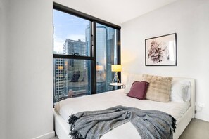 A hotel room with a large window, a bed with a white headboard, a lamp, and a framed picture on the wall. - Collins St CBD escape| Pool, Gym, Sauna & Parking (Melbourne)