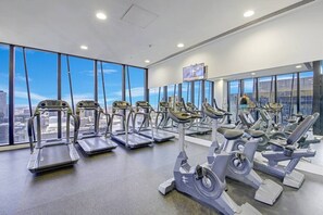 A gym with treadmills and elliptical machines, large windows with a city view, and a flat-screen TV on the wall. - Collins St CBD escape| Pool, Gym, Sauna & Parking (Melbourne)