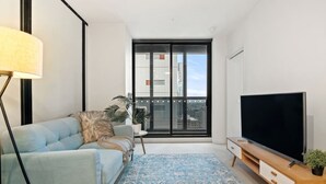 En moderne stue med sofa, fjernsyn, gulvlampe og et stort vindue med udsigt over byen.