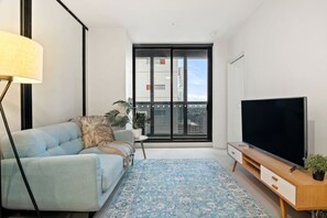 A modern living room with a sofa, a television, a floor lamp, and a large window with a city view. - Collins St CBD escape| Pool, Gym, Sauna & Parking (Melbourne)