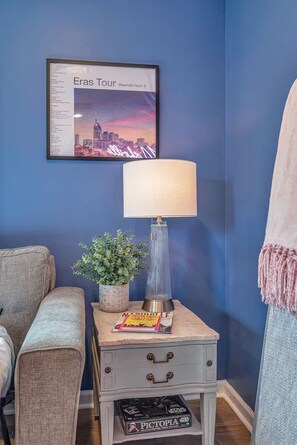 A room with a blue wall, a framed picture of a cityscape, a lamp, a small table with a plant, a magazine, and a Star Wars box. - The Swiftie: Sleeps 5, Pool, City & River Views, Near Broadway (Nashville)