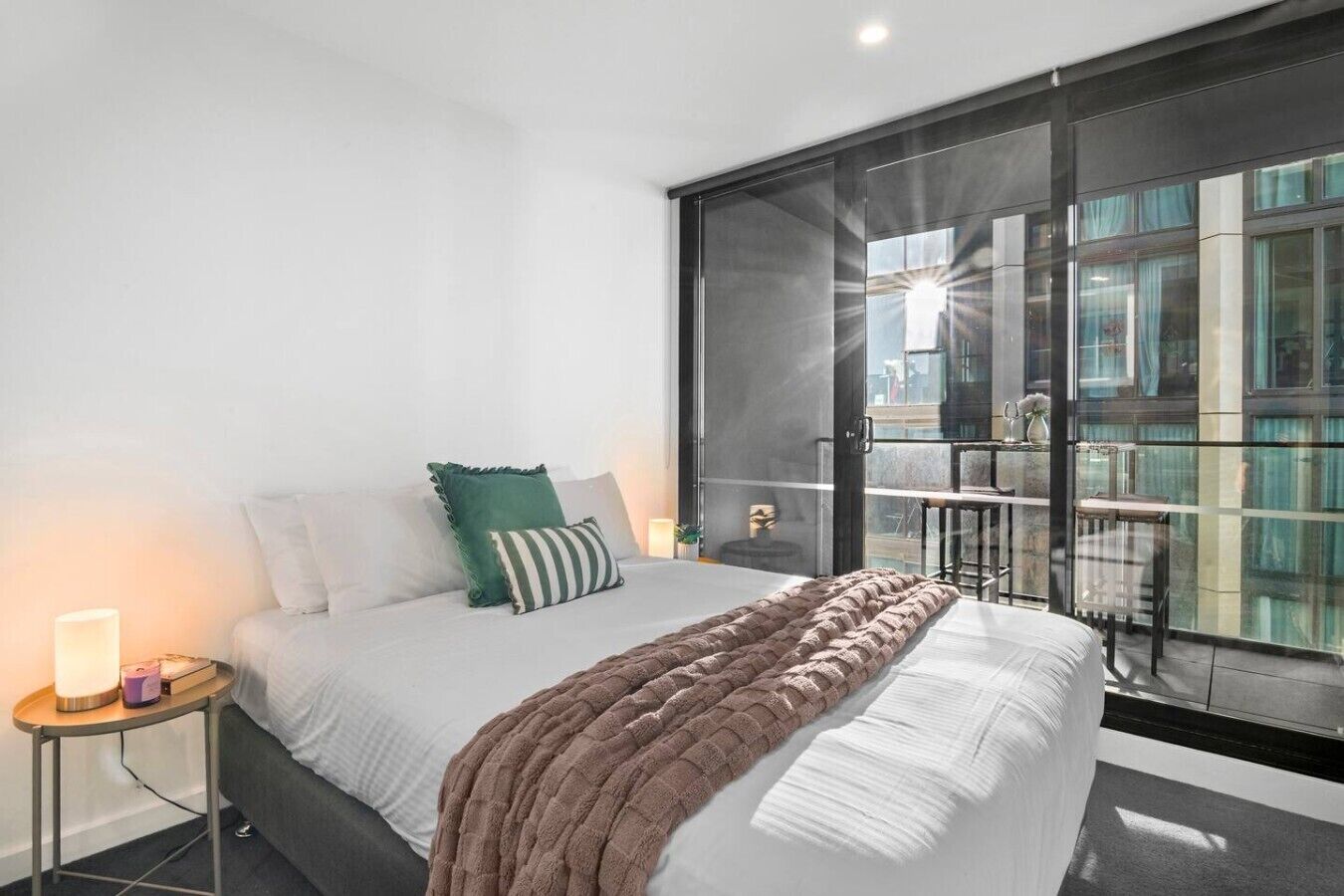 A modern bedroom with a large bed, a bedside table with a lamp, and a view of a balcony with a table and chairs.