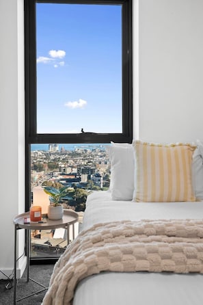 A bedroom with a large window, a bed with white and striped pillows, a bedside table with a candle and a plant, and a view of a cityscape. - International Southbank 2BR2Bath1Car Jacuzzi +more (Southbank)