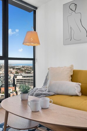 Ein Zimmer mit großem Fenster, einer Couch mit Kissen, einem Tisch mit zwei Tassen und einer Pendelleuchte.