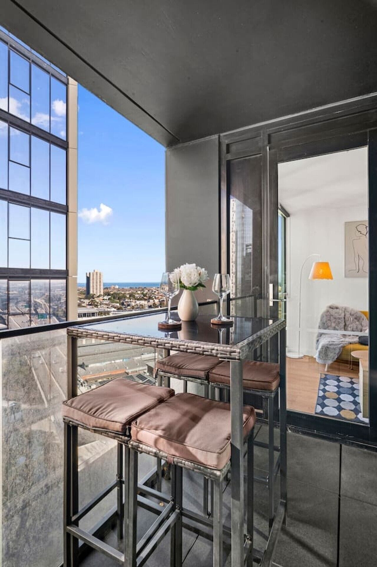 A balcony with a glass barrier, a table with a vase and glasses, and a view of the cityscape.