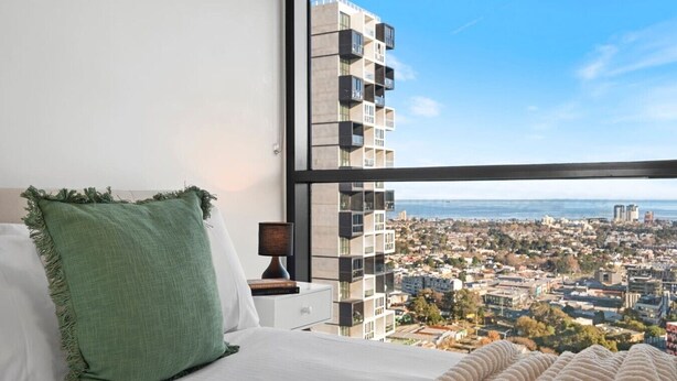 Quarto com uma janela grande, uma cama, mesa de cabeceira com abajur e vista para a cidade.