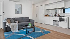 Uma sala de estar moderna com um sofá cinza, uma mesa de centro e um tapete azul. Ao lado, encontra-se uma cozinha com armários brancos, um fogão e um micro-ondas.