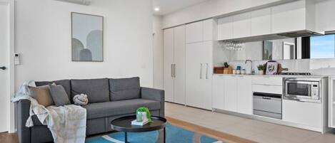 A modern living room with a grey sofa, a coffee table, and a blue rug. Adjacent is a kitchen with white cabinets, a stove, and a microwave.
