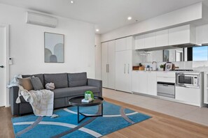 Ein modernes Wohnzimmer mit einem grauen Sofa, einem Couchtisch und einem blauen Teppich. Daneben befindet sich eine Küche mit weißen Schränken, einem Herd und einer Mikrowelle.
