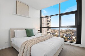 Een slaapkamer met een groot raam, een bed met witte lakens en uitzicht op een stadsgezicht.